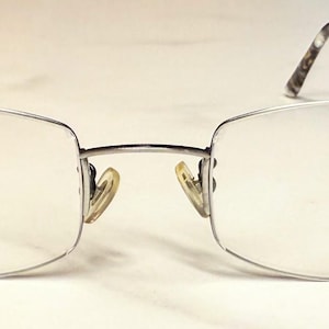 Puede incluir: Un par de gafas con montura de metal plateado y lentes transparentes. Las patillas tienen un diseño estampado. Las almohadillas nasales son amarillas. Las gafas están sobre una superficie blanca.