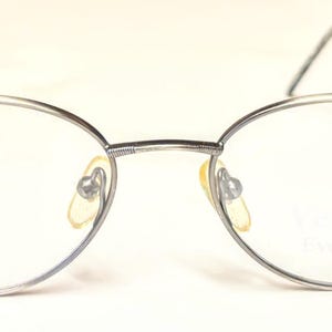 Può includere: Occhiali con montatura argentata e lenti ovali. Le montature hanno un design sottile e delicato con un motivo sottile sulle aste. I naselli sono di colore giallo chiaro. Le lenti sono trasparenti.