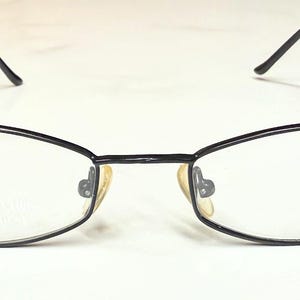 May include: Black-framed eyeglasses with clear lenses. The glasses have a rectangular shape with rounded edges and a thin metal frame. The nose pads are a light beige color. The temples are thin and extend to the ear pieces.