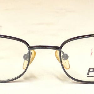 May include: A pair of eyeglasses with a dark gray metal frame and oval lenses. The brand name "Pezzo" is printed on the right lens. The glasses have a classic, understated design.