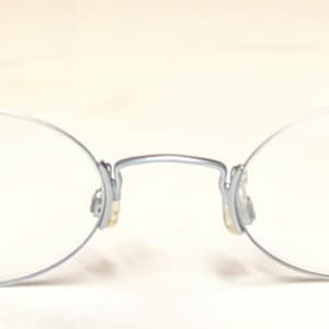 May include: Oval-shaped eyeglasses with a silver metal frame and black temples. The lenses are clear, and the brand name "EYETEL" is printed on the right lens. The glasses have a minimalist design.