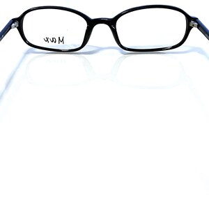 K&ouml;nnte beinhalten: Schwarze Brille mit ovalen Gl&auml;sern und einem schwarzen Rahmen. Die Brille hat ein modernes Design mit d&uuml;nnen B&uuml;geln. Der Text "49 M" ist auf dem Glas sichtbar. Die Brille ist vor einem wei&szlig;en Hintergrund platziert.