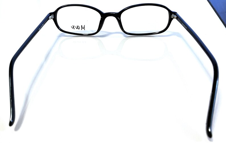 K&ouml;nnte beinhalten: Schwarze Brille mit ovalen Gl&auml;sern und einem schwarzen Rahmen. Die Brille hat ein modernes Design mit d&uuml;nnen B&uuml;geln. Der Text "49 M" ist auf dem Glas sichtbar. Die Brille ist vor einem wei&szlig;en Hintergrund platziert.