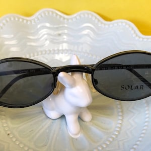 Puede incluir: Gafas de sol ovaladas negras con lentes oscuros. Las gafas tienen el texto "SOLAR" impreso en la lente derecha y "Design Italy" impreso en la lente izquierda.