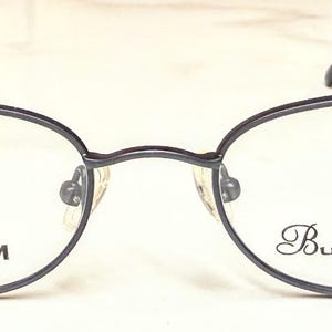 May include: Dark grey oval titanium eyeglasses with slender frames. The left lens features the word "TITANIUM", and the right lens has the text "Burns & Allen". The arms are thin and extend to the ear pieces.