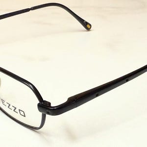 May include: Black rectangular eyeglasses with the brand name "PEZZO" printed on the lens. The frames are black with a thin, sleek design. The temples are also black and have a slight curve for a comfortable fit.