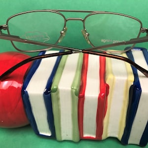 Może przedstawiać: Srebrne okulary w stylu lotniczym z brązowymi zausznikami spoczywające na stosie kolorowych książek. Okulary mają przezroczyste szkła, a na prawej zauszniku nadrukowano napis "Spring Hinge".