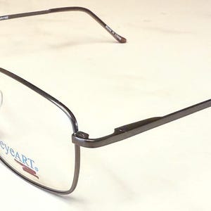 Pode incluir: Um par de óculos com armação de metal cinza escuro. As lentes são transparentes e a armação tem formato retangular. A marca "eyeART" é visível na lente inferior esquerda.