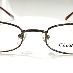 Op de afbeelding: Bruine metalen bril met heldere glazen. De merknaam "CLUB MAD" staat op het rechter glas gedrukt.