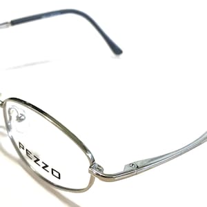 May include: Silver-toned metal eyeglasses with a black rubber temple tip. The glasses have a clear lens and the brand name "PEZZO" is printed on the right lens.