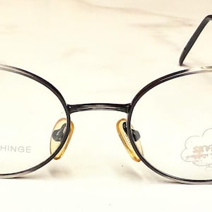 Könnte beinhalten: Ovale Brille mit einem dunkelgrauen Metallrahmen. Die Gläser sind klar, und die Bügel sind dünn und schwarz. Auf einem Glas steht "SPRING HINGE". Die Brille hat ein kleines, goldfarbenes Nasenpolster.
