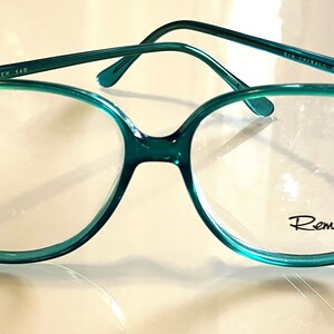 Könnte beinhalten: Türkise Kunststoff-Brille mit klaren Gläsern. Der Markenname "Remi" ist auf dem rechten Glas aufgedruckt.