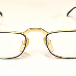 Op de afbeelding: Goudkleurige bril met zwarte pootjes en heldere glazen. De rechthoekige monturen hebben een gouden brug en het woord "STUDIO" staat op het rechterglas. De bril staat tegen een witte achtergrond.