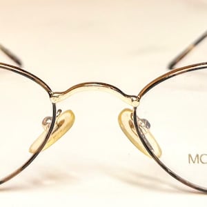 May include: Oval eyeglasses with a gold-coloured metal frame. The lenses are clear, and the nose pads are a light yellow. The text "MODA ITALIA" is printed on the right lens. The arms of the glasses are thin and dark.