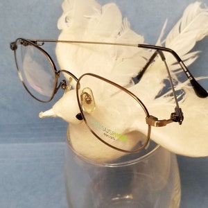 Puede incluir: Gafas con montura dorada y diseño de doble puente. Las lentes son transparentes y las monturas tienen un aspecto fino y similar a un alambre. La marca "MATSUSHIMA" es visible en la montura. Las gafas están sobre un fondo de plumas blancas.