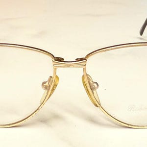 May include: Gold-toned eyeglasses with a semi-rimless design. The frames have a curved shape with a bridge and nose pads. The temples are thin and black. The lenses are clear, and the text "Roberto Elite" is visible on the lens.
