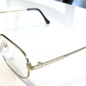 May include: Gold-rimmed eyeglasses with a rectangular lens shape. The frames have a delicate, thin design with a decorative pattern on the temples. The earpieces are black. The glasses are set against a white background.