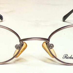May include: Oval-shaped eyeglasses with a thin, brown metal frame. The lenses are clear, and the nose pads are a light yellow. The brand name "Roberto Elliot" is printed on the right lens. The glasses have a small red sticker on the left lens.