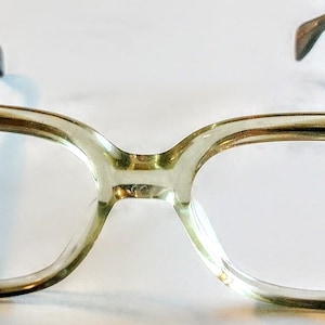 Op de afbeelding: Een vintage bril met een licht olijfgroen montuur. De glazen zijn helder en de pootjes zijn donkerder van kleur. De bril heeft een klassiek, retro design.