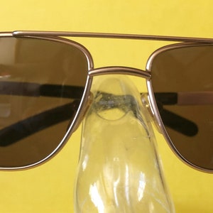May include: A pair of gold-toned aviator sunglasses with brown lenses. The sunglasses are sitting on a clear plastic stand.