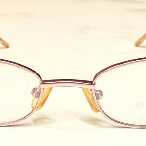 May include: A pair of oval-shaped eyeglasses with a light pink metal frame. The lenses are clear, and the temples are a light brown color. The glasses have a delicate, minimalist design.