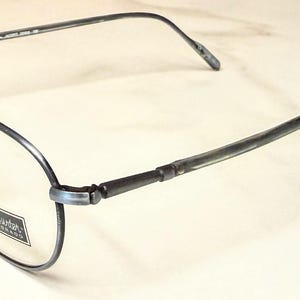 May include: Oval-shaped eyeglasses with a dark gray metal frame. The lenses are clear, and the nose pads are a light tan color. The temples are thin and extend back to the ear pieces. The brand name is visible on the lens.