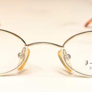 May include: Oval spectacles with a gold-coloured frame and brown temples. The clear lenses feature the text "J-VISION EYEWEAR" on the right lens. These glasses are designed for vision correction.