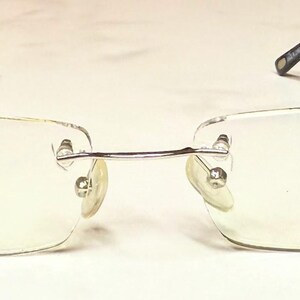 May include: Rimless eyeglasses with rectangular lenses. The glasses have a silver bridge and temples with dark brown arms. The text "STAINLESS STEEL HAND MADE ACETATE" is printed on the left lens, and the word "RAVE" is printed on the right lens.