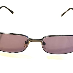 Pode incluir: Óculos de sol retangulares com armação de metal cinza escuro e lentes roxas. Design clássico e minimalista. As hastes finas estendem-se até às extremidades.