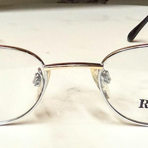 May include: Oval-shaped eyeglasses with a gold-colored frame and dark brown temples. The lenses are clear, and the text "ROKKO" is printed on one of the lenses. The glasses are designed for vision correction.