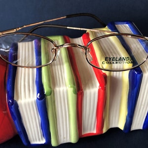 May include: Gold-tone metal eyeglasses with a rectangular frame and clear lenses. The glasses are resting on a stack of colorful books with the words "Eyelands Collection" printed on the side of the books.