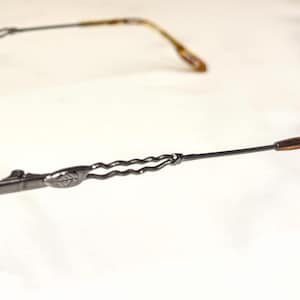May include: A pair of eyeglasses with a dark gray metal frame. The round lenses are held in place by a thin frame. The temples have a decorative twisted design and end with a tortoiseshell pattern.