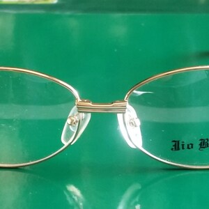 Könnte beinhalten: Ovale Brille mit goldfarbenem Rahmen, schwarzem Kunststoffgestell und dem Schriftzug "Jio Bendini" auf der rechten Linse.