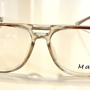 Może przedstawiać: Okulary z przezroczystego plastiku z brązowymi akcentami i napisem "Max" nadrukowanym na prawej soczewce.