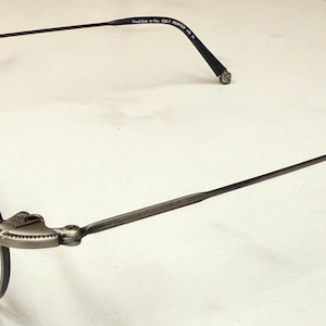 May include: A pair of eyeglasses with a gunmetal gray frame and round lenses. The temples are thin and black, with a small decorative element at the end. The frame has a unique hinge design.