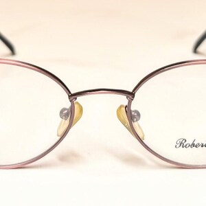 May include: Oval-shaped eyeglasses with a mauve frame and black temples. The lenses are clear, and the nose pads are yellow. The text "Roberto Elliot" is printed on the right lens. A blue dot is on the right lens.