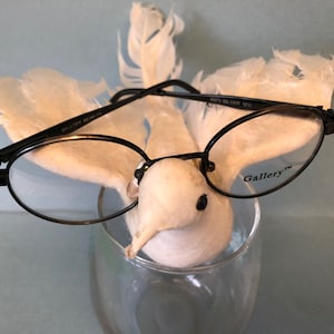 May include: Black round glasses with the word "Gallery" printed on the right lens. The glasses are perched on a white feathery bird figurine.