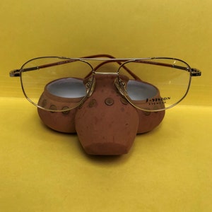 May include: Gold-rimmed aviator glasses with clear lenses resting on a brown ceramic holder. The holder has a textured surface and the glasses have the text "J-VISION EYEWEAR" printed on the right lens.