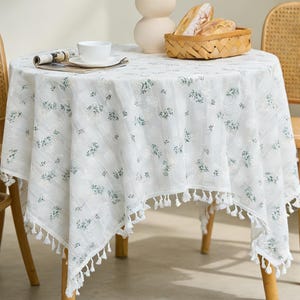 Pode incluir: Uma toalha de mesa redonda branca com estampa floral e acabamento de borlas cobre uma pequena mesa de madeira. Um vaso de flores, uma cesta de pão e uma xícara e pires estão sobre a mesa. Duas cadeiras de vime estão ao lado da mesa.