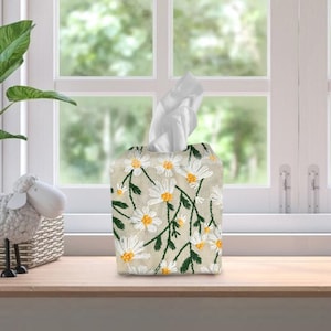 May include: A white tissue box cover with a floral design featuring white daisies with yellow centres and green stems on a beige background.