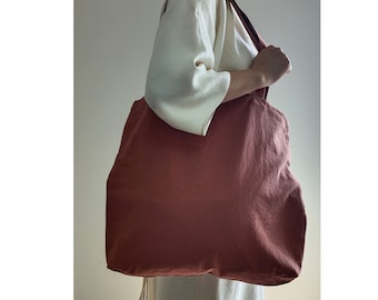 May include: A large, rust-colored tote bag made of canvas with a single long strap. The bag is being held by a person wearing a white shirt.