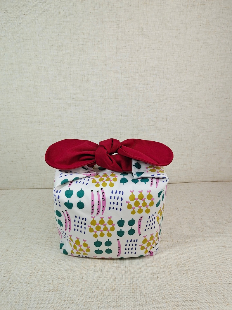 May include: A white fabric gift bag with a red ribbon tied around it. The bag has a colorful pattern of green leaves, pink and yellow flowers, and white dots.