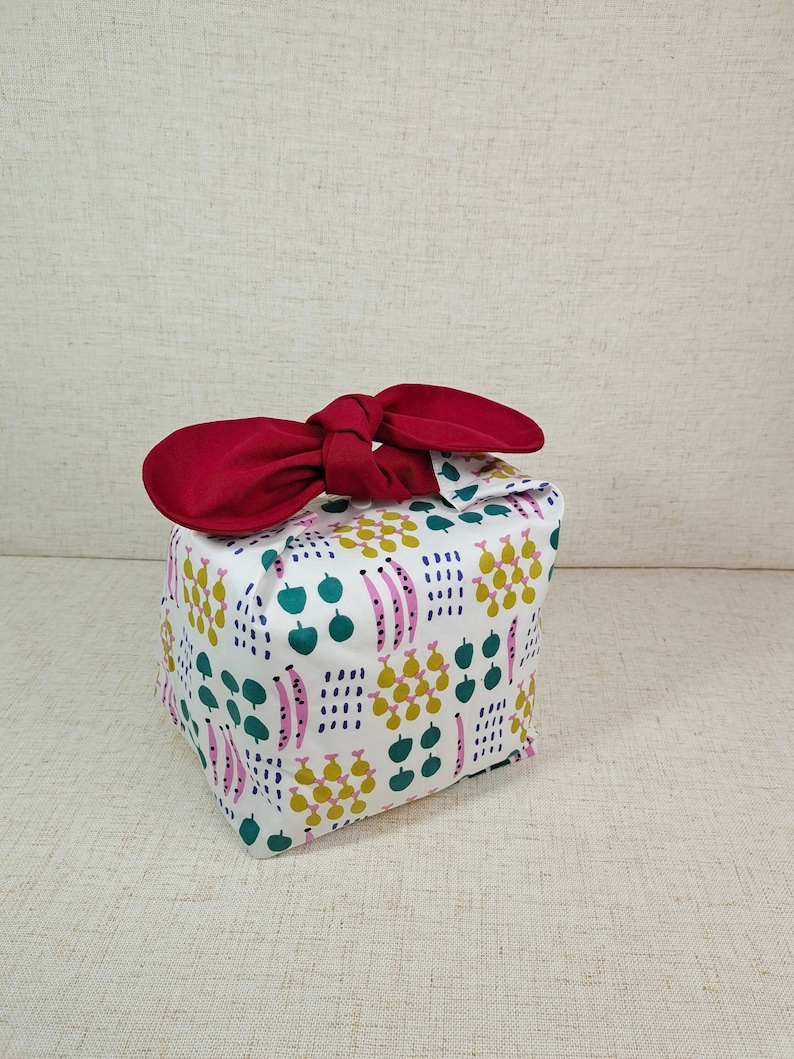 May include: A white fabric gift bag with a red bow. The bag has a pattern of green leaves, pink and yellow pears, and pink and white stripes.