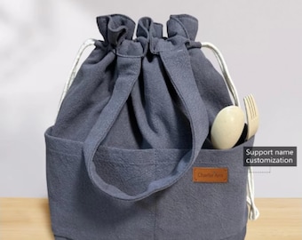 Puede incluir: Una bolsa de almuerzo de lona gris con cierre de cordón y dos bolsillos. La bolsa tiene una etiqueta de cuero marrón con el texto "Charlie Ann" en ella. La bolsa está sentada sobre una superficie de madera. Soporte de personalización de nombre.