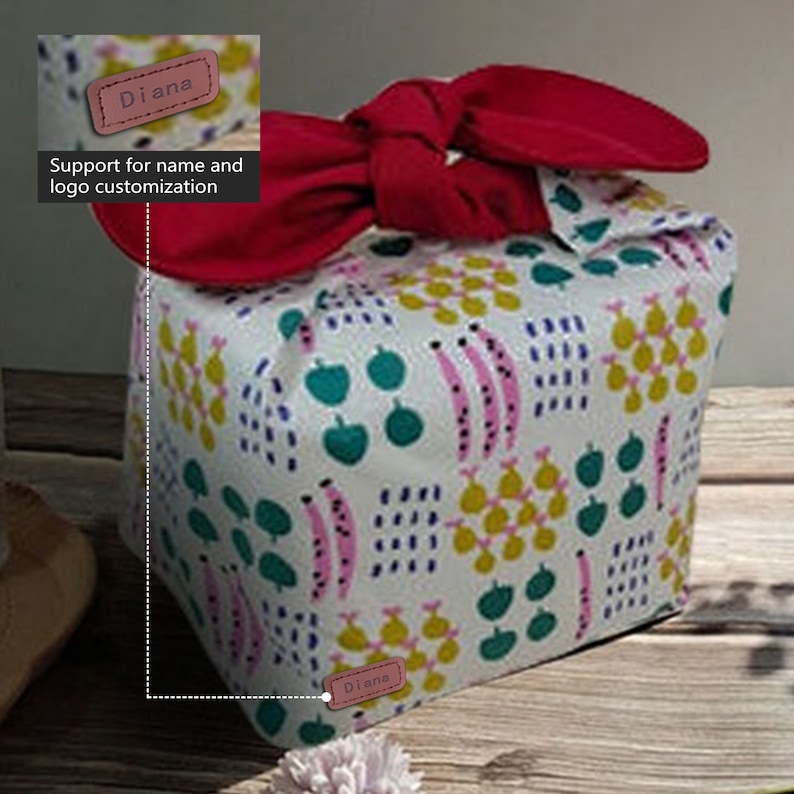 May include: A white fabric gift bag with a red bow and a geometric pattern of green, pink, and yellow shapes. The bag has a brown leather tag with the name "Diana" on it. Support for name and logo customization.