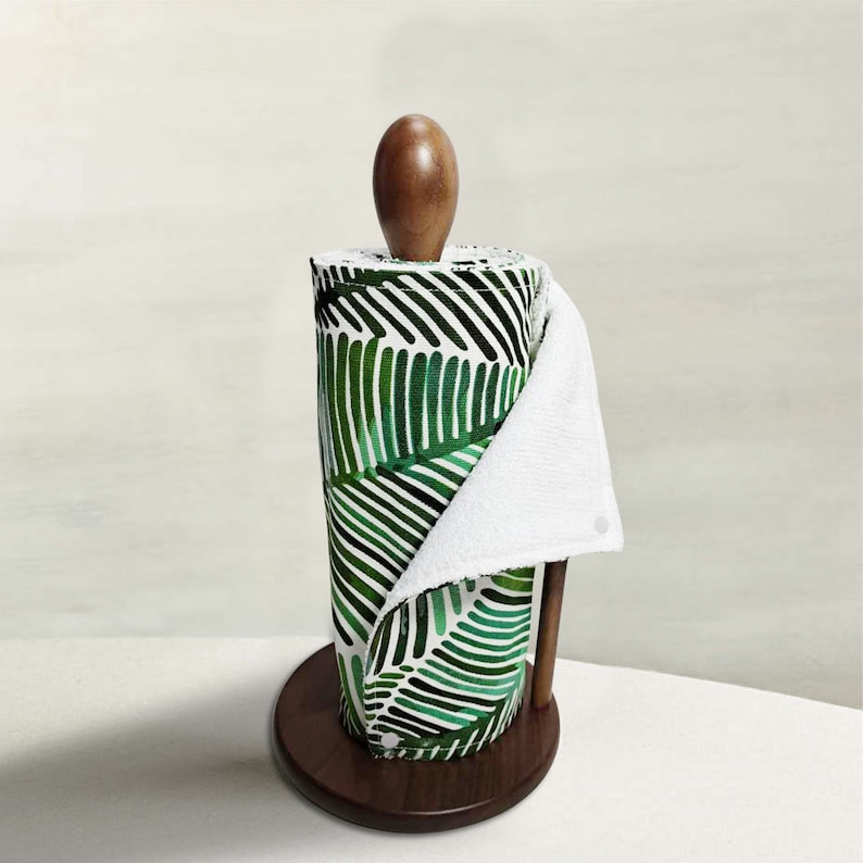Pu&ograve; includere: Un porta rotoli di carta da cucina in legno con un rotolo di carta da cucina a motivi verdi e bianchi. La carta da cucina ha un design a foglie tropicali.