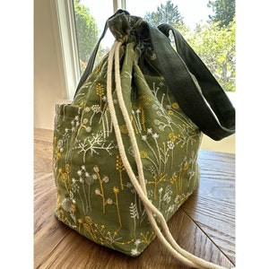 May include: A green canvas tote bag with embroidered white and yellow flowers. The bag has a drawstring closure and two long handles.