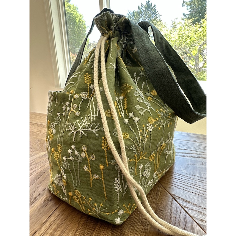May include: A green canvas tote bag with embroidered white and yellow flowers. The bag has a drawstring closure and two long handles.