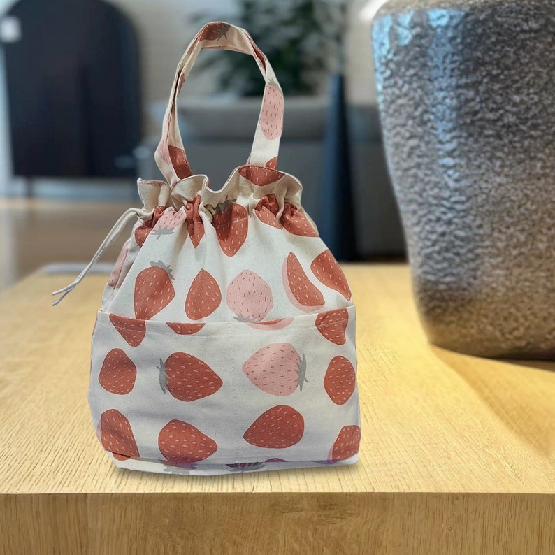 May include: A white canvas drawstring bag with a pink and red strawberry print. The bag has a single handle and a drawstring closure.
