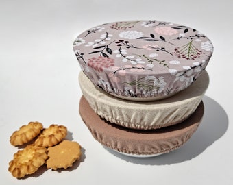 May include: Three reusable food covers in a stack. The top cover is a light gray with a floral pattern in shades of pink, white, and brown. The middle cover is a beige color and the bottom cover is a light brown. The covers are made of fabric and are designed to fit over bowls or plates to keep food fresh.